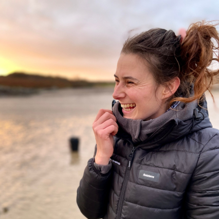 Lucy is standing on the beach with the sun setting over the horizon. She is wearing a black Project Seagrass coat.